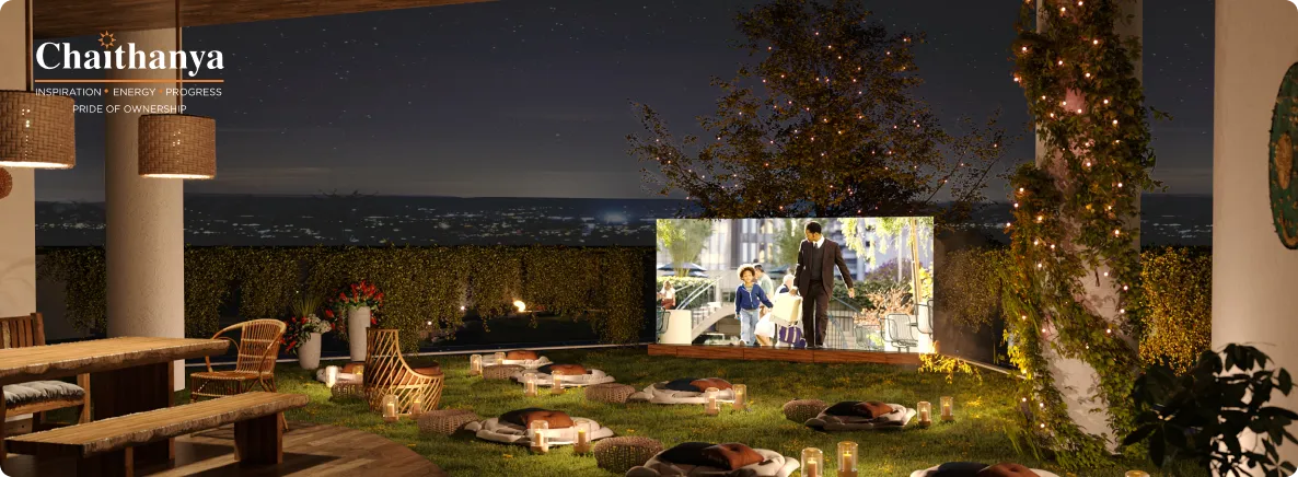 Outdoor nighttime Chaithanya event space with lanterns, cushions on the grass, and a large projection screen showing people; city lights in the distance.