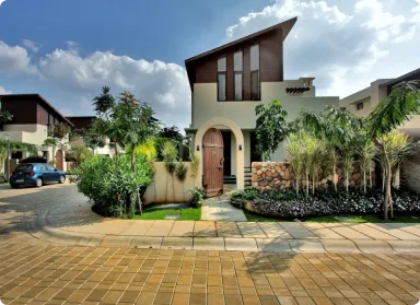 Modern two‑story house with an arched front entry, tall angled roof, and tropical landscaping; curved driveway with a blue car on the left side.