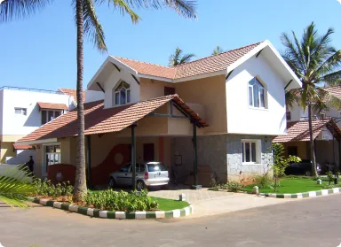 Two-story house with white and beige exterior, a red-tiled roof, a covered carport with a silver car, and palm trees against a blue sky.