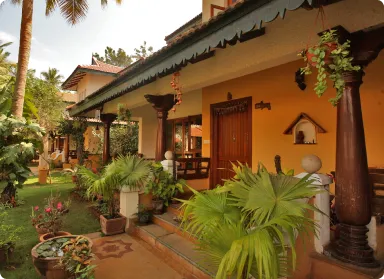 Sunlit tropical porch of a yellow-orange house with dark wooden columns, hanging plants, and potted palms along a tiled path.