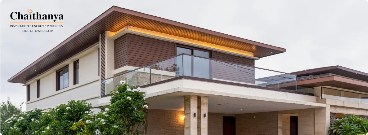 Modern two-story house with glass balcony railing, warm exterior lights, and landscaped bushes; Chaithanya logo in the top-left corner.