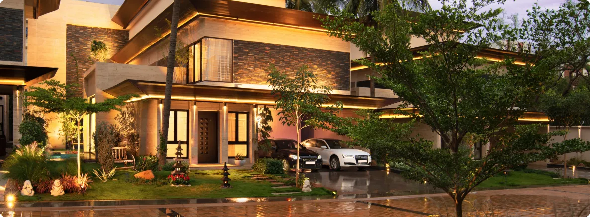 Luxurious two-story house with warm exterior lighting, stone accents, palm trees, and two cars parked in the driveway at dusk.