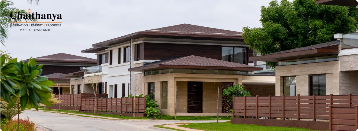Modern two-story houses with brown roofs and beige stone facades along a quiet street, Chanthanya logo in the corner.