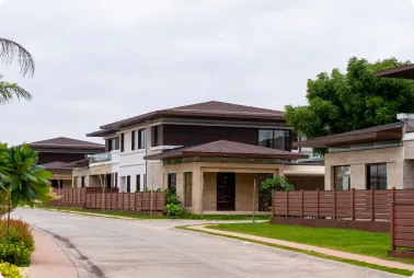 Quiet suburban street with modern two-story houses behind brown wooden fences and trimmed lawns under an overcast sky.
