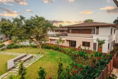 Modern two-story white house with wooden accents, surrounded by a manicured lawn, trees, and flower beds in a fenced yard at sunset (exterior shot).