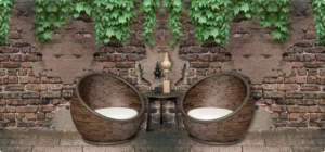 Two round wicker chairs with white cushions face a small round table on a stone patio, against a brick wall draped with ivy.