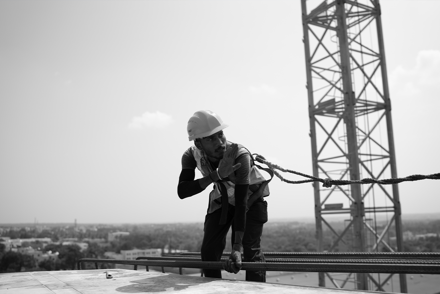Construction worker in a hard hat and safety vest crouches on a rooftop, holding a rope attached to a cable barrier with a tall metal structure in the background.